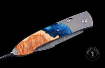 Folding knife featuring a handle of natural burl wood and sparkling blue resin, with a grey titanium frame, a Damascus steel blade, and a black braided lanyard.