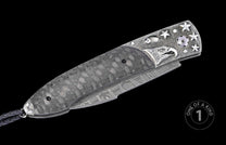 Folding knife with grey carbon fiber handle, engraved silver eagle head, stars, Damascus steel blade, and black braided lanyard.