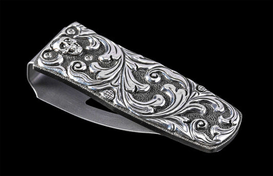 Silver metal money clip with an engraved skull, intricate scrollwork, and textured background, viewed at an angle.