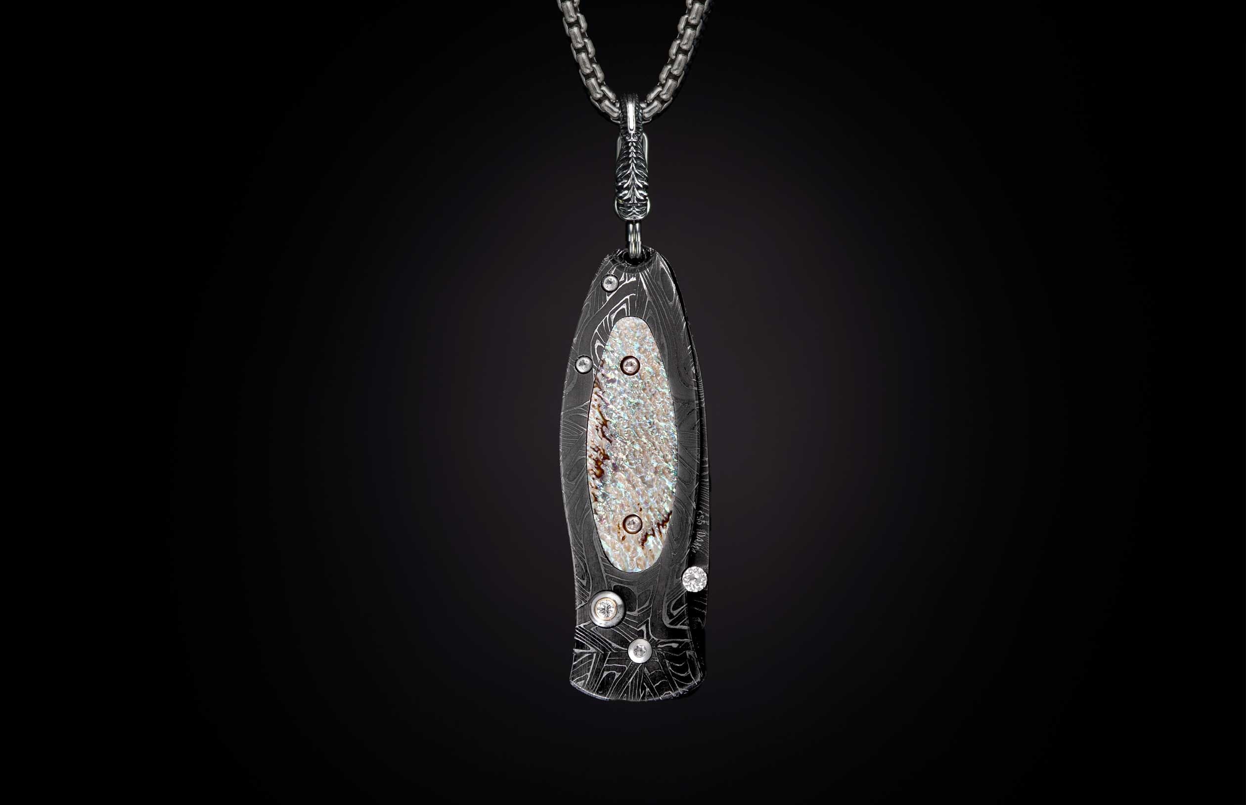 Stylized dark metal knife pendant with wavy Damascus pattern, iridescent mother-of-pearl inlay, and clear stones on a silver chain.