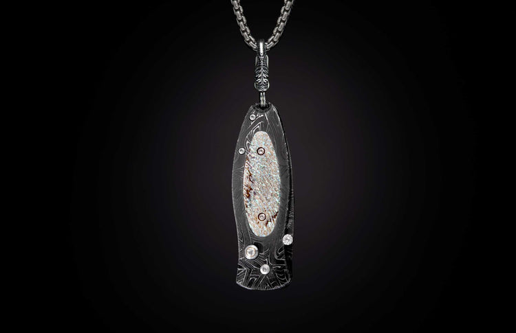 Stylized dark metal knife pendant with wavy Damascus pattern, iridescent mother-of-pearl inlay, and clear stones on a silver chain.