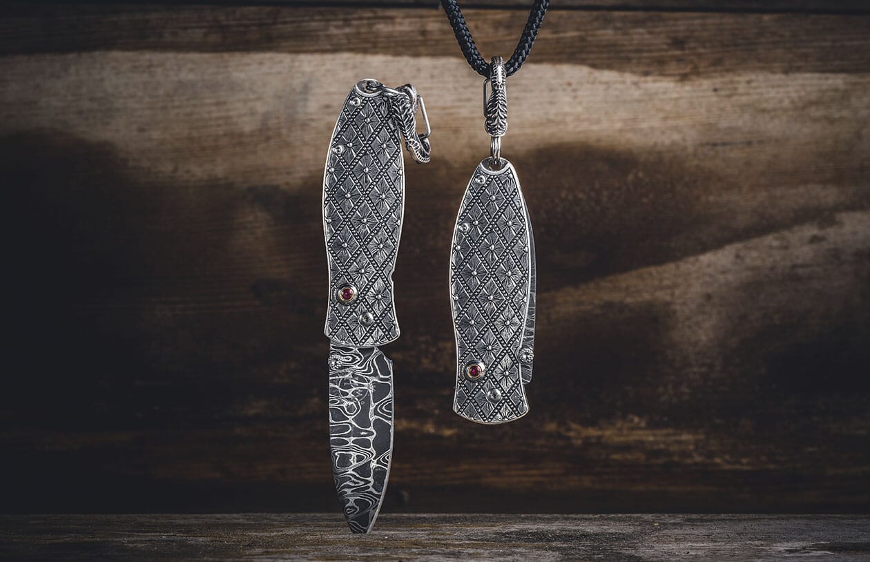 Two ornate silver-toned folding knives with red gemstones and patterned Damascus blades; one open, one closed hanging.