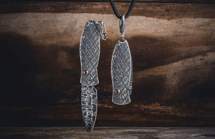 Two ornate silver-toned folding knives with red gemstones and patterned Damascus blades; one open, one closed hanging.