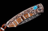 Ornate folding knife with engraved silver handle, blue stone, banded brown and white material, and a patterned blade.