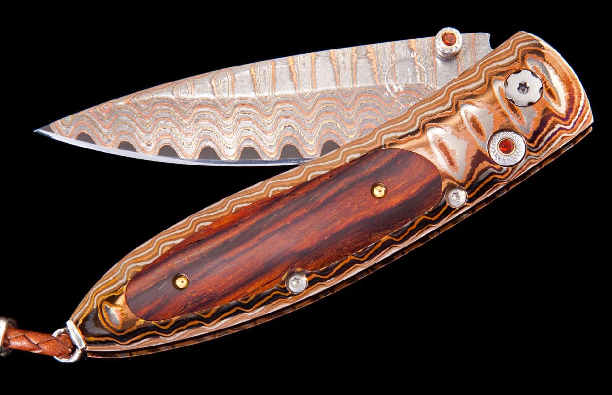 Open folding knife with wavy Damascus steel blade and matching layered metal handle, featuring wood inlay and red gems.