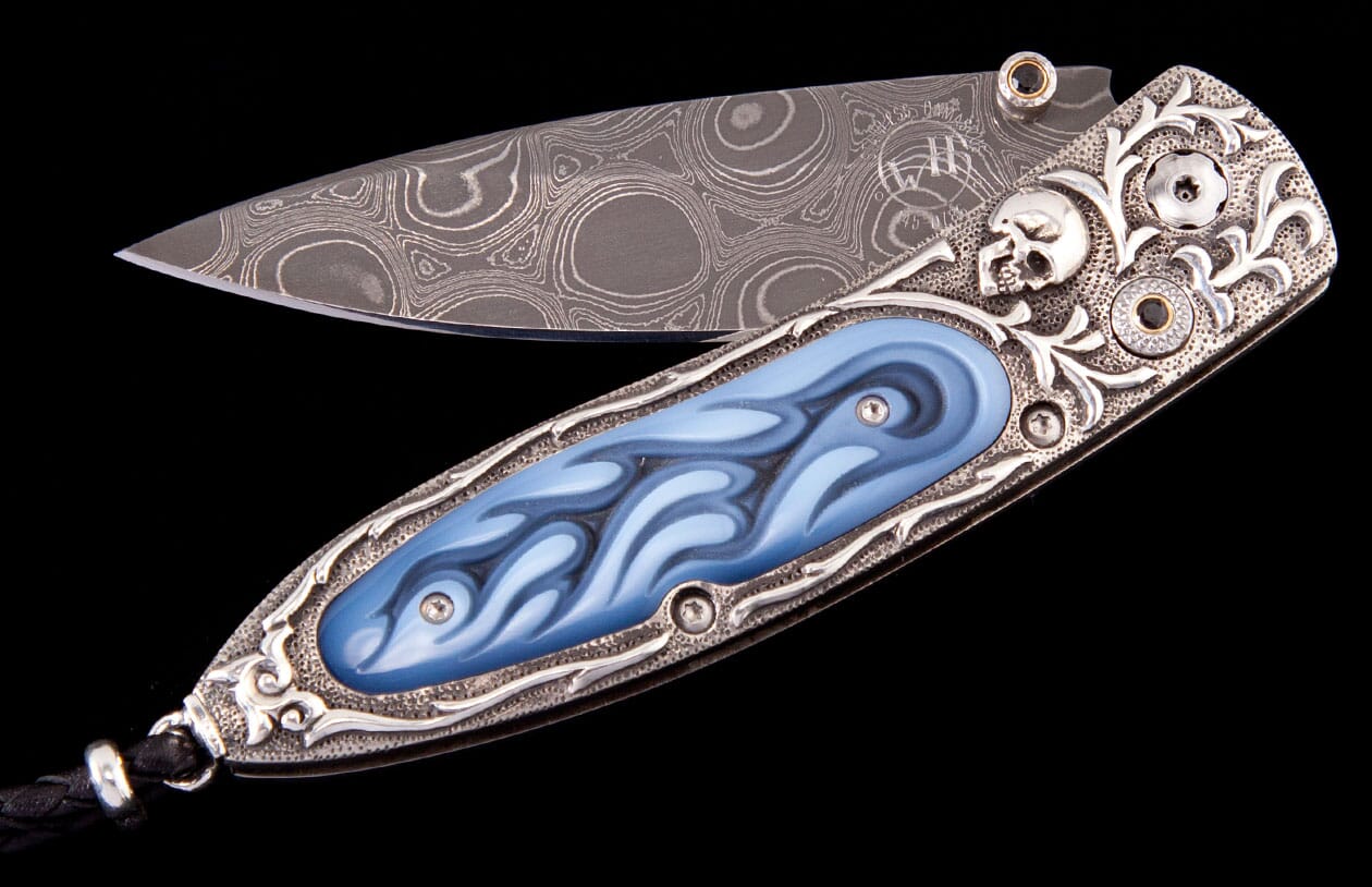 A custom pocket knife with an open Damascus blade and an ornate silver handle featuring a skull and blue flame inlay.