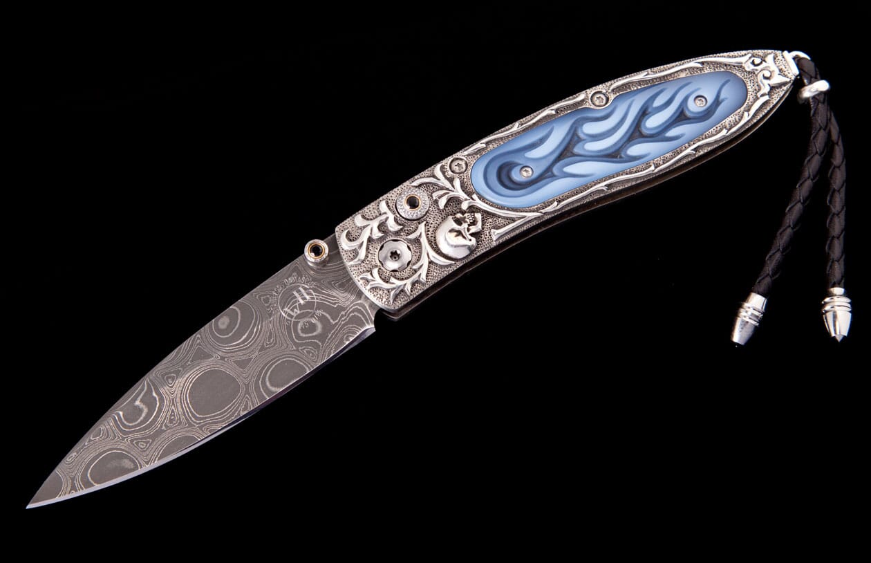 Ornate silver folding knife with a blue flaming skull design, a Damascus blade, and a black leather lanyard.