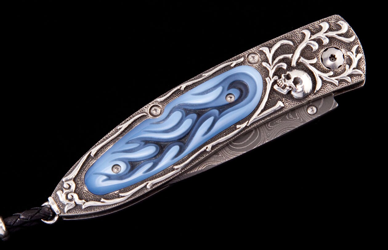 A silver custom folding knife with engraved skull and floral patterns, blue flame-like inlay, and a Damascus blade.