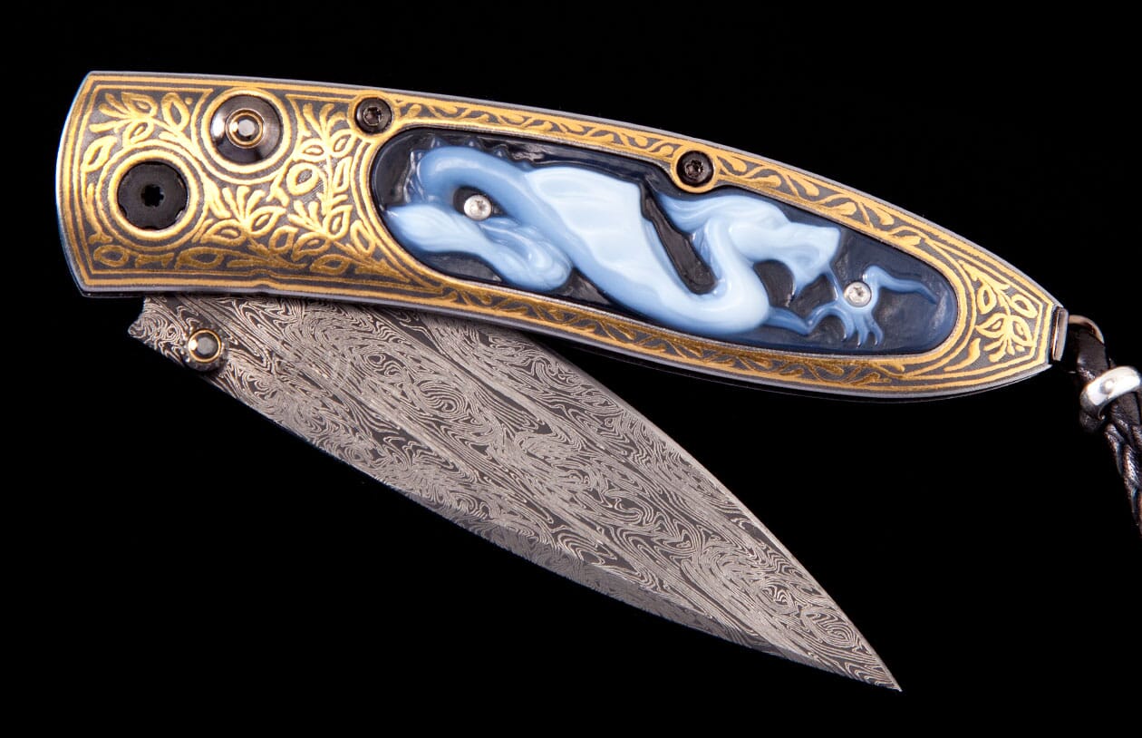 Ornate folding knife with a gold engraved handle featuring a carved blue dragon inlay with gems, and a Damascus steel blade.