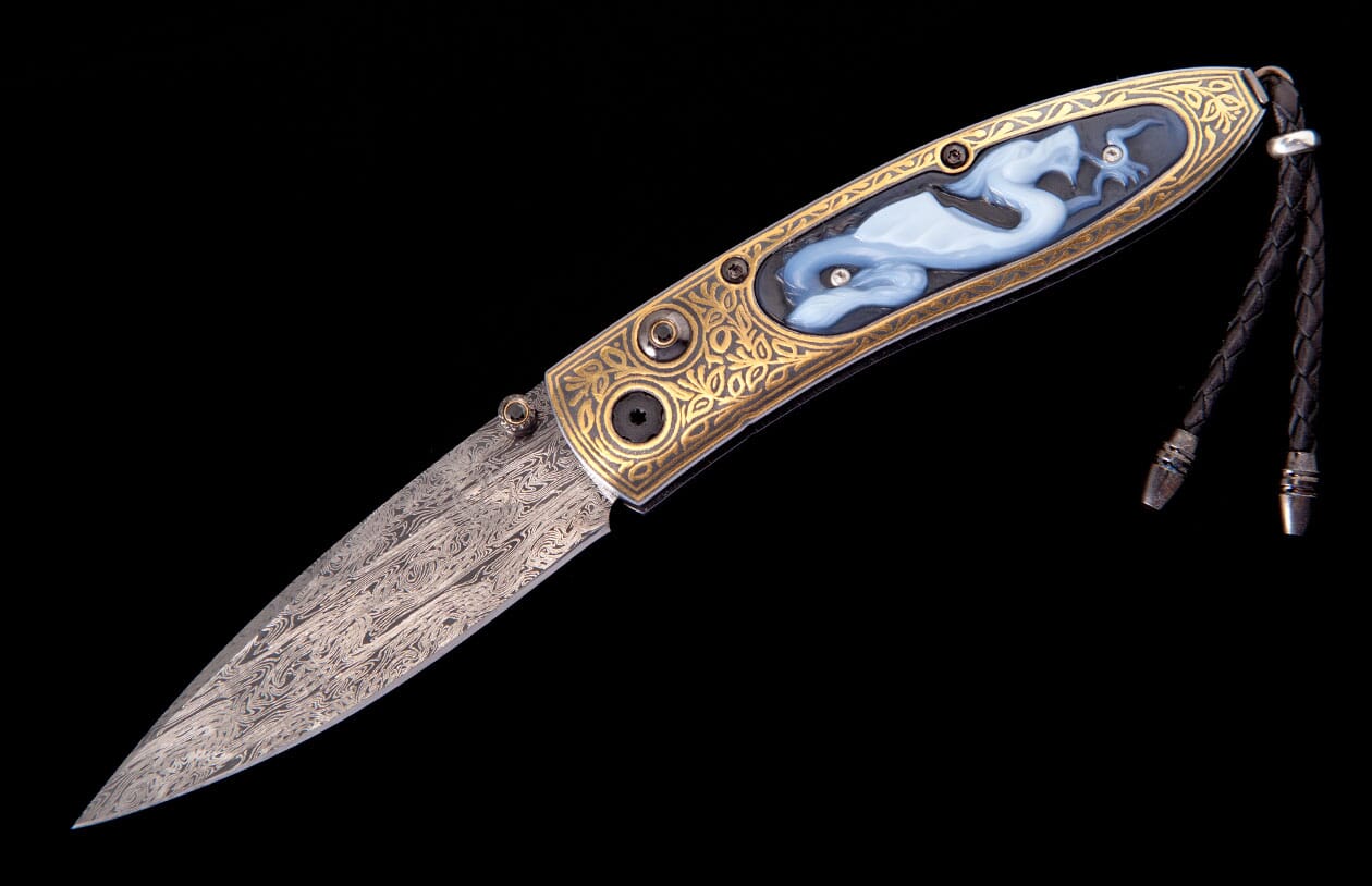 Gold-handled knife with a damascus blade, intricate scrolling, a blue carved dragon inlay, and a black braided lanyard.