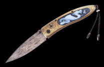 Gold-handled knife with a damascus blade, intricate scrolling, a blue carved dragon inlay, and a black braided lanyard.
