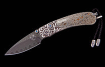 Ornate folding knife with a wavy Damascus steel blade, a textured bone handle, and silver scrollwork featuring blue gemstones, with a braided black lanyard.