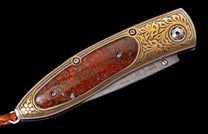Ornate folding knife with a red patterned handle, gold scrollwork, a Damascus steel blade, and a leather lanyard.