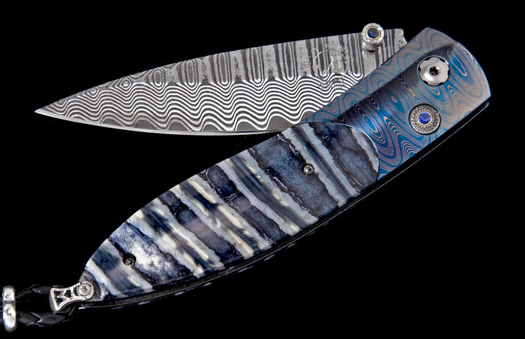 Partially open knife with a wavy Damascus blade, blue textured metal and striped scales, blue gems, and a black braided lanyard.
