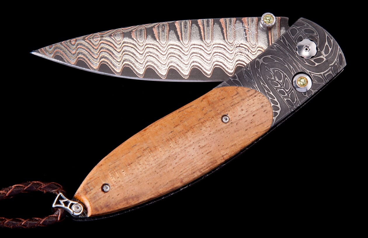 Folding knife with wavy Damascus steel blade and light brown wood handle by Hildgund, featuring an etched bolster, yellow gemstones, and braided leather lanyard.