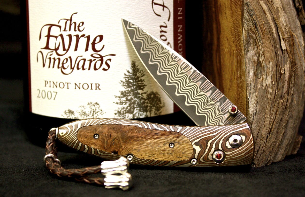 Damascus steel folding knife with a wavy pattern blade, a wooden inlay handle, red accents, and a braided leather strap.