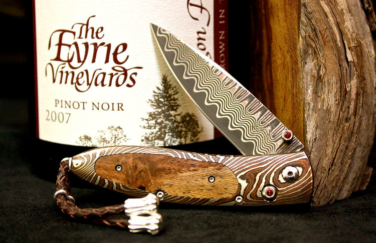 Damascus steel folding knife with a wavy pattern blade, a wooden inlay handle, red accents, and a braided leather strap.