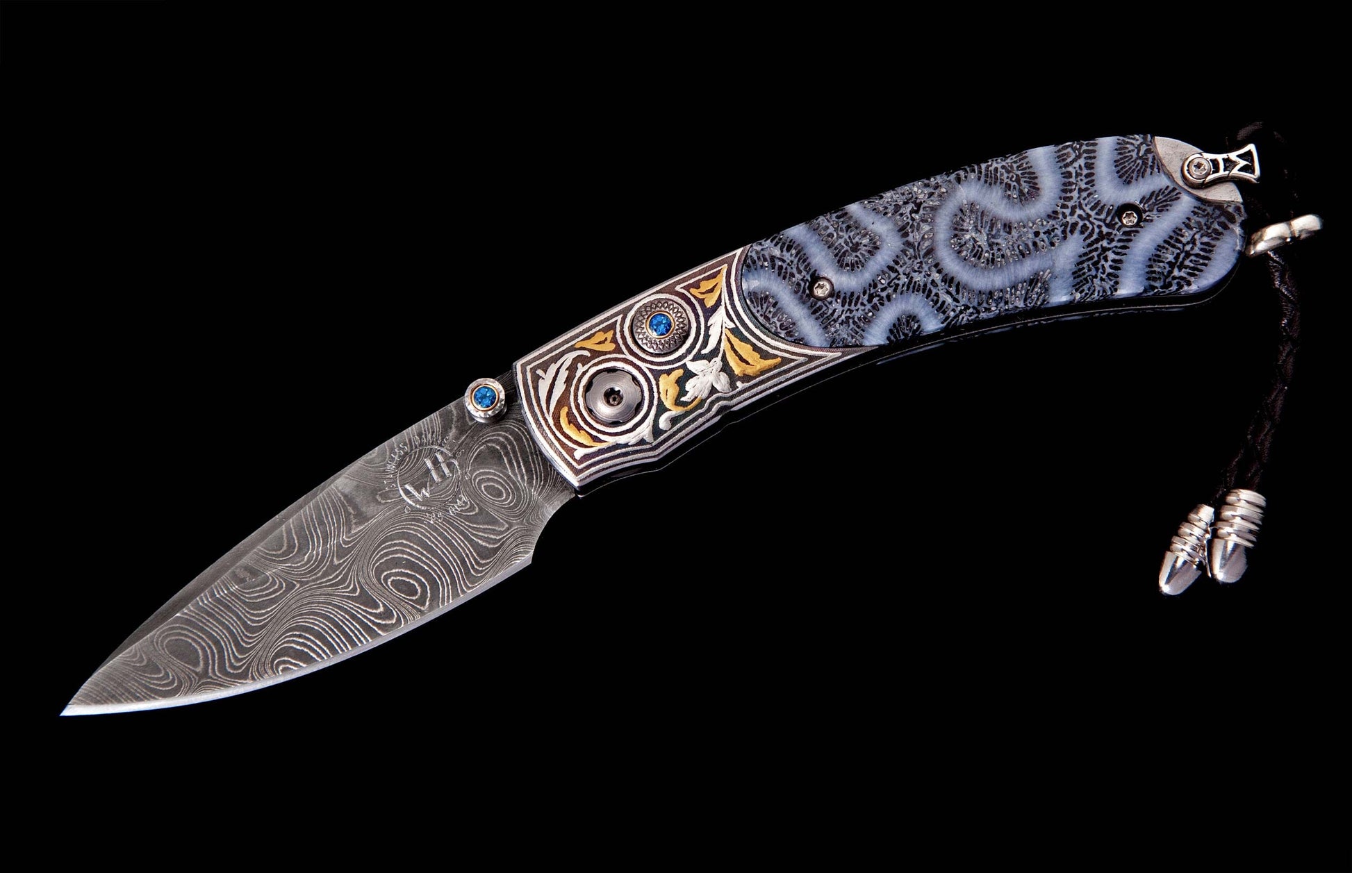 Folding knife with a swirling Damascus steel blade, ornate silver and gold-tone bolsters, blue gemstones, unique fossilized coral handle, and a black leather lanyard.