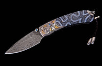 Folding knife with a swirling Damascus steel blade, ornate silver and gold-tone bolsters, blue gemstones, unique fossilized coral handle, and a black leather lanyard.
