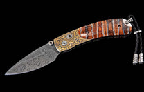Folding knife with a swirling Damascus blade, ornate gold-engraved bolster, and striped brown handle with a black braided lanyard.