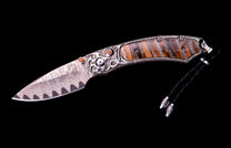 Ornate knife with a wavy Damascus steel blade, a silver handle featuring scrollwork, orange inlays, striped brown panels, and a black braided lanyard with silver beads.