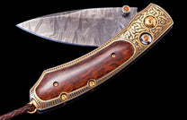 Ornate folding knife with Damascus steel blade, gold-etched handle, dark wood inlay, orange gems, and braided leather lanyard.