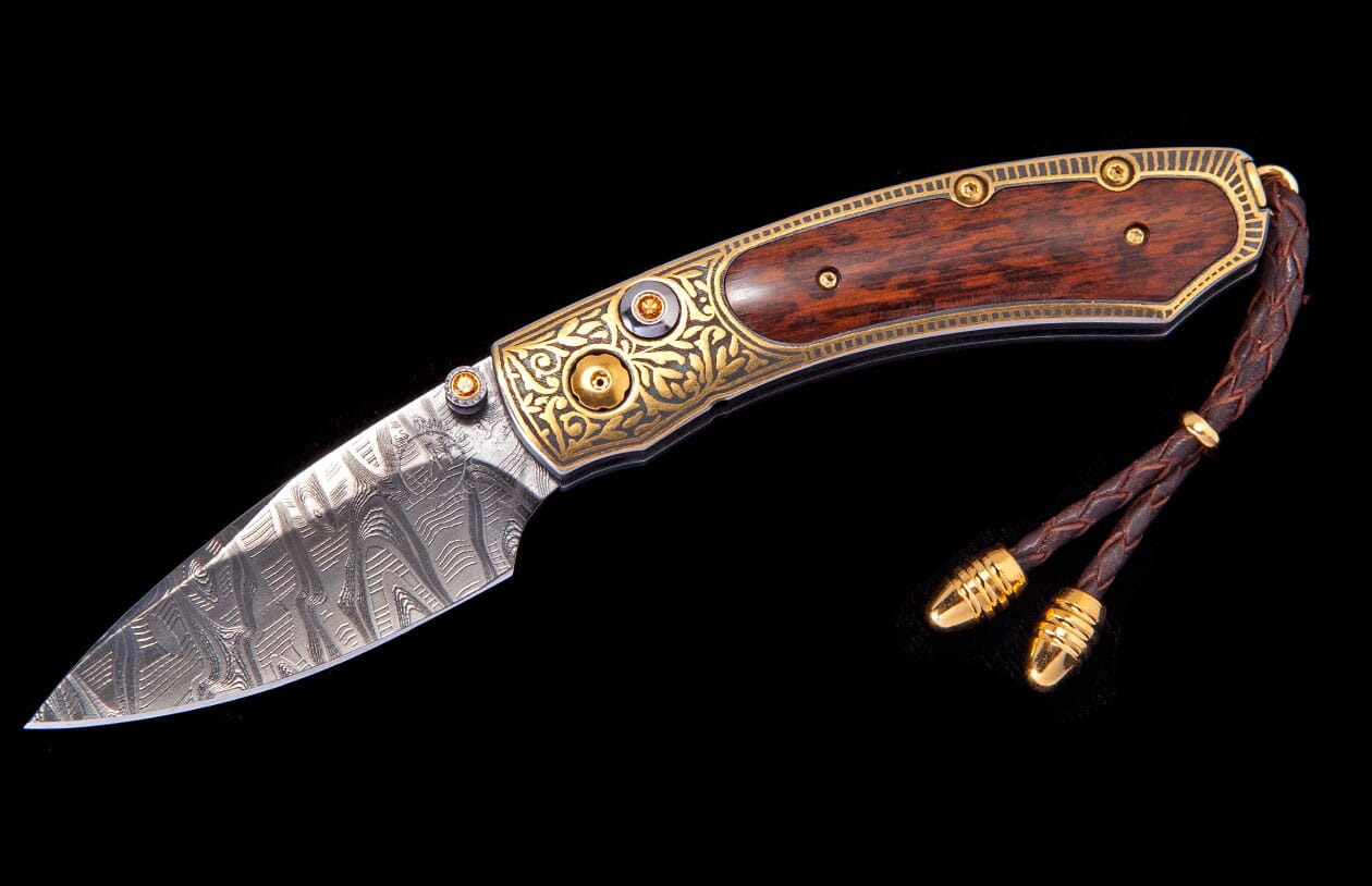Luxury folding knife with damascus blade, dark wood handle, ornate gold-colored bolsters, and braided leather lanyard.
