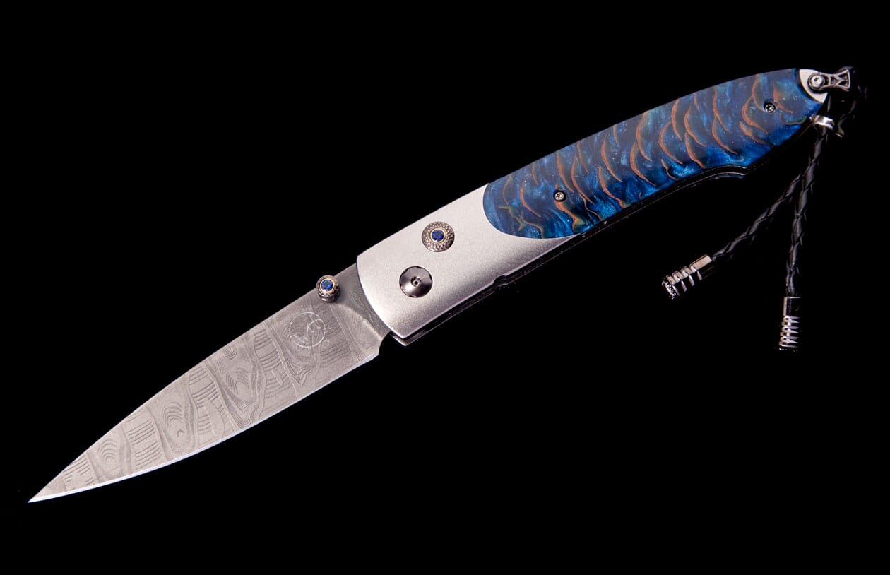 A custom folding knife featuring a patterned Damascus blade, a blue and orange scale handle with blue gems, and a black leather lanyard.