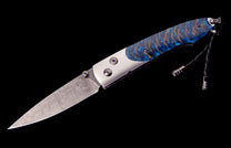 A custom folding knife featuring a patterned Damascus blade, a blue and orange scale handle with blue gems, and a black leather lanyard.