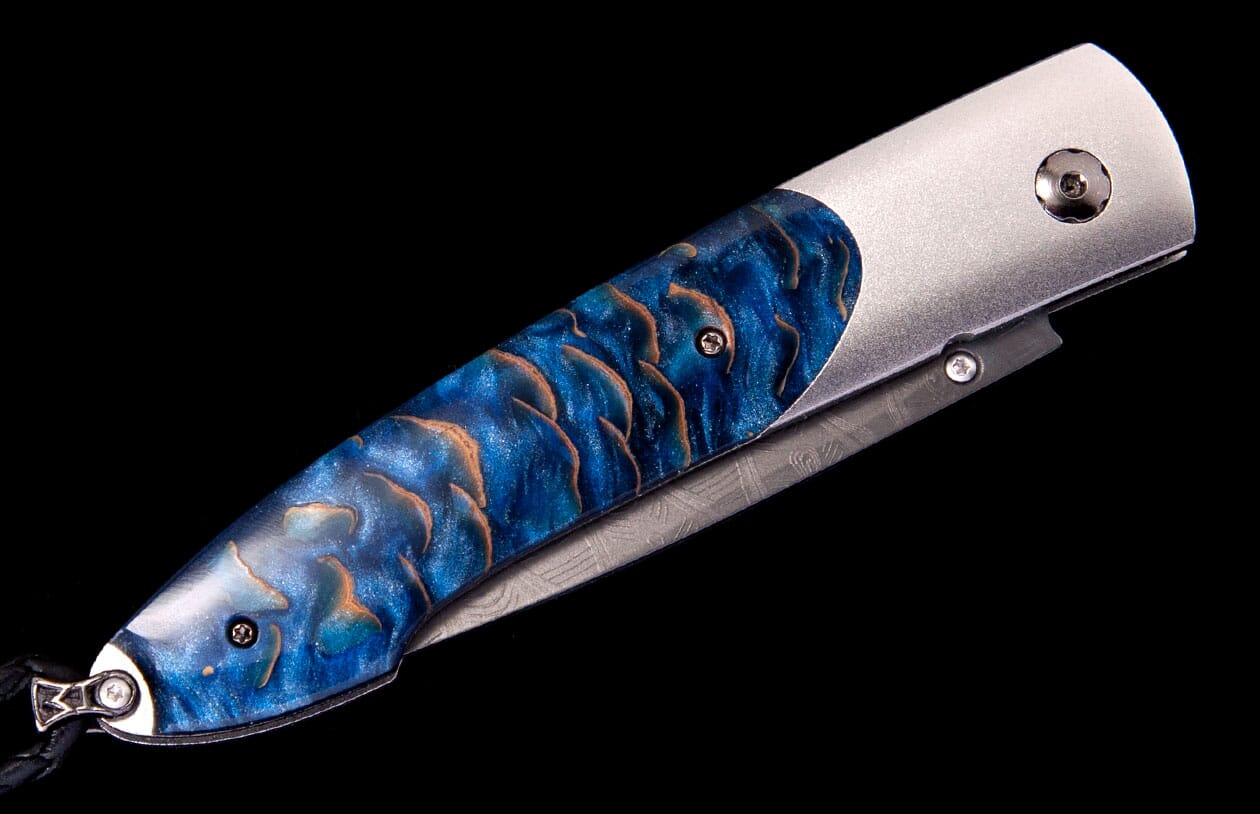 A folding knife with a blue and copper scale-patterned handle, brushed silver bolster, and a visible Damascus blade.