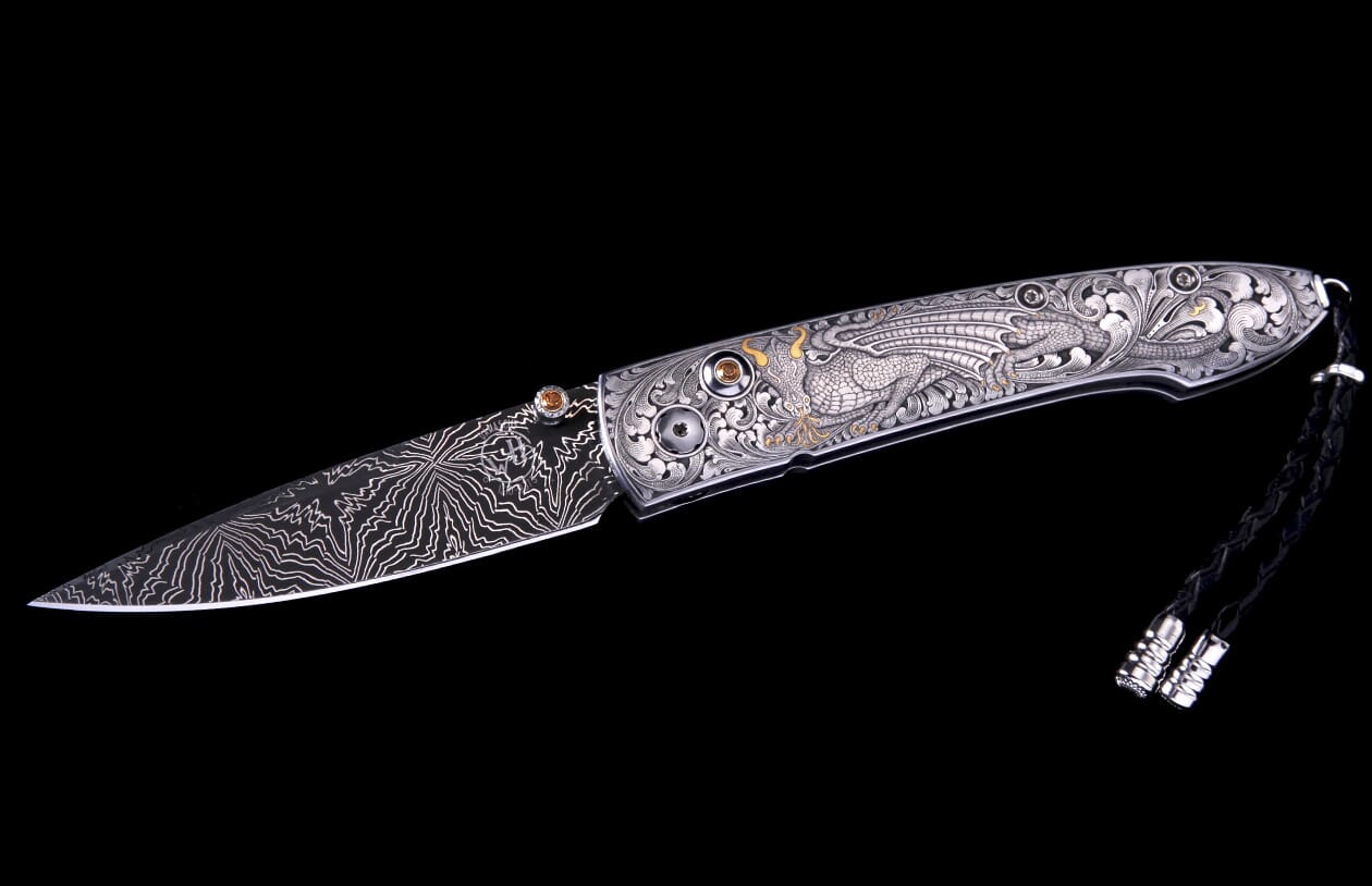 An elaborately engraved folding knife with a patterned Damascus blade, silver handle featuring a dragon, gold accents, amber gems, and a black lanyard.