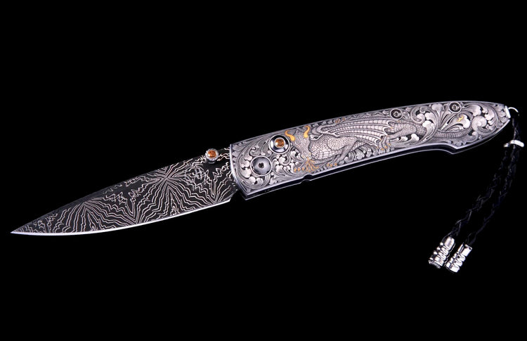 An elaborately engraved folding knife with a patterned Damascus blade, silver handle featuring a dragon, gold accents, amber gems, and a black lanyard.
