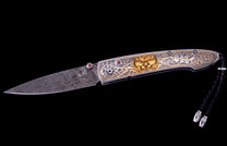 Ornate custom folding knife featuring a swirling Damascus steel blade, a gold owl, and silver scroll engraving on the handle, with red gemstone details and a black braided lanyard.