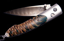 Partially open folding knife with a wavy patterned blade and a textured, scale-like handle in teal, gold, and brown.