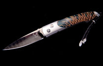 Folding knife with a wavy patterned blade, a silver-toned body, and a teal and brown scale-textured handle with a black braided lanyard.