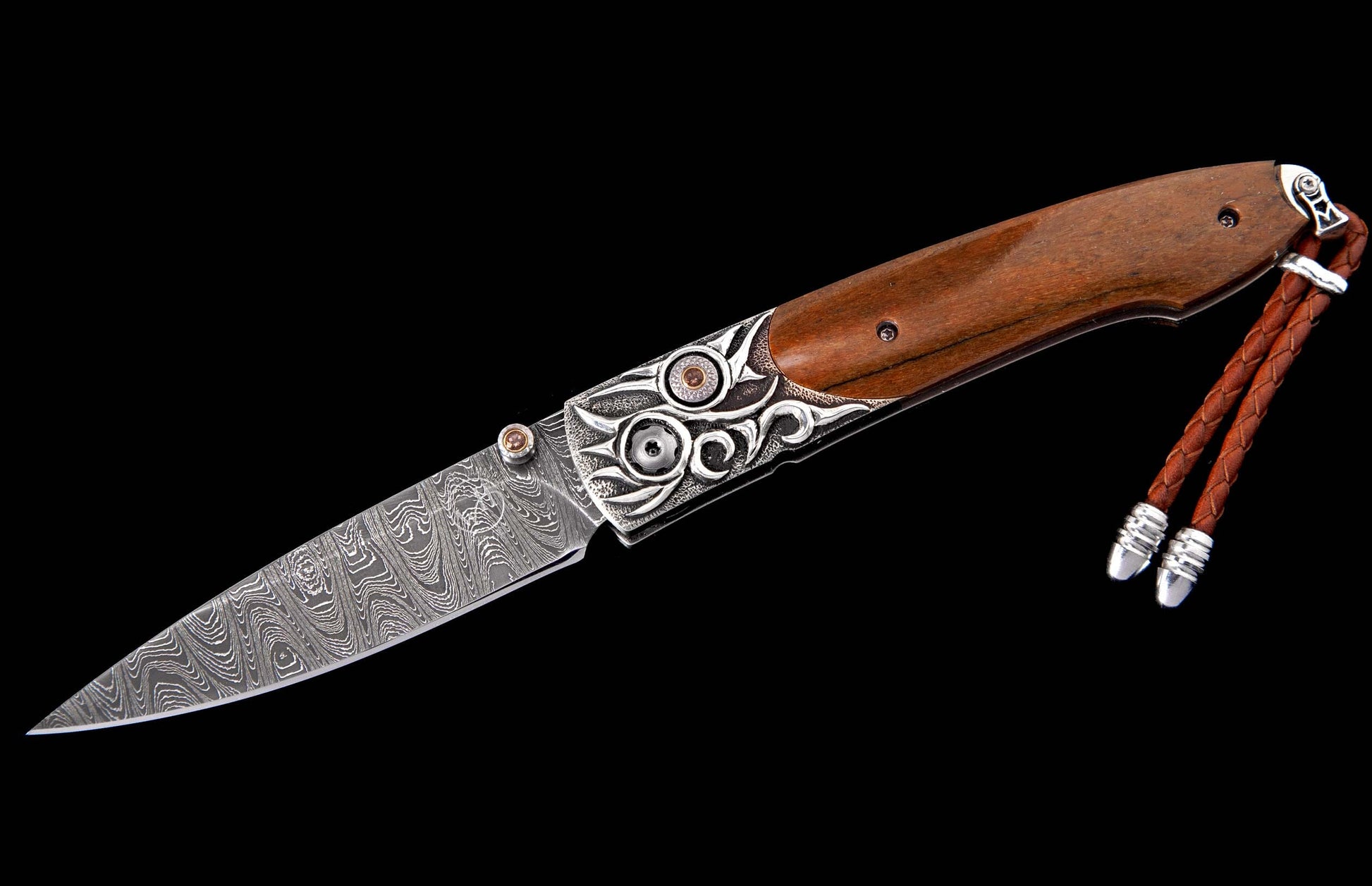 Folding knife with Damascus steel blade, ornate silver bolster with gems, brown wooden handle, and braided leather lanyard.