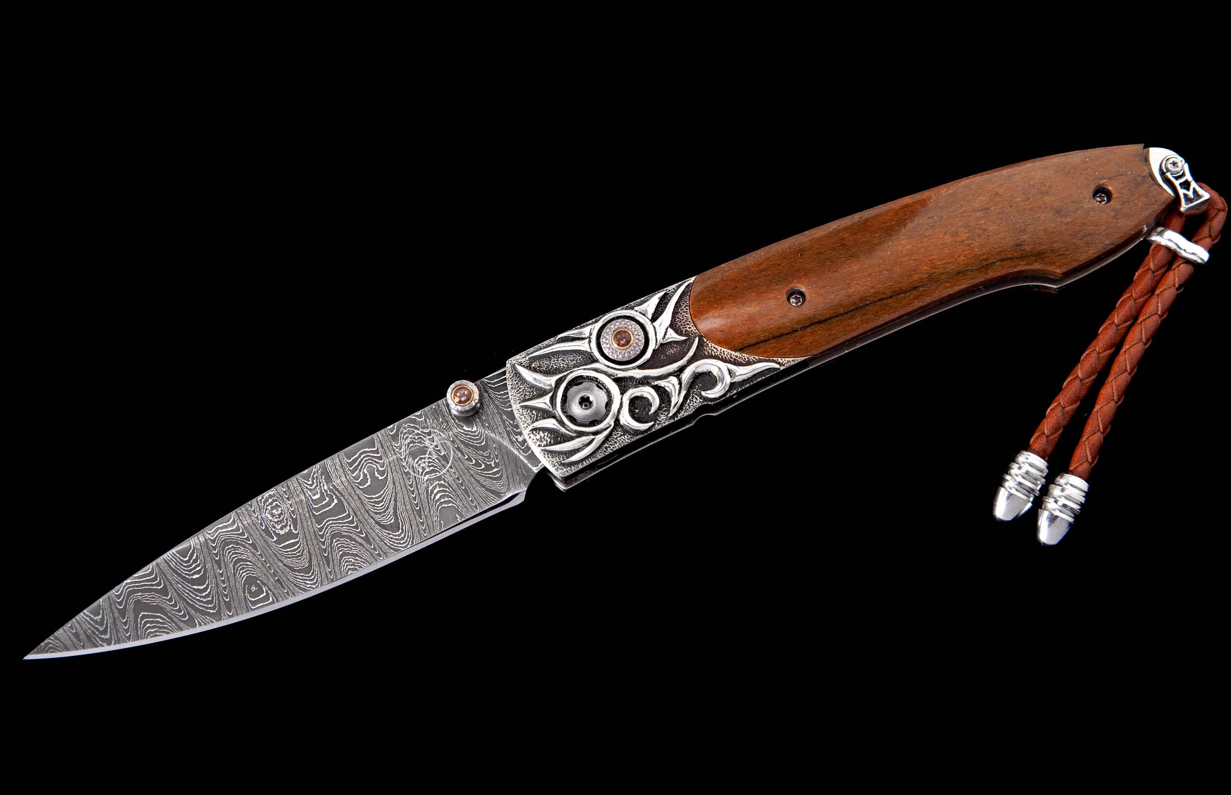 Folding knife with Damascus steel blade, ornate silver bolster with gems, brown wooden handle, and braided leather lanyard.