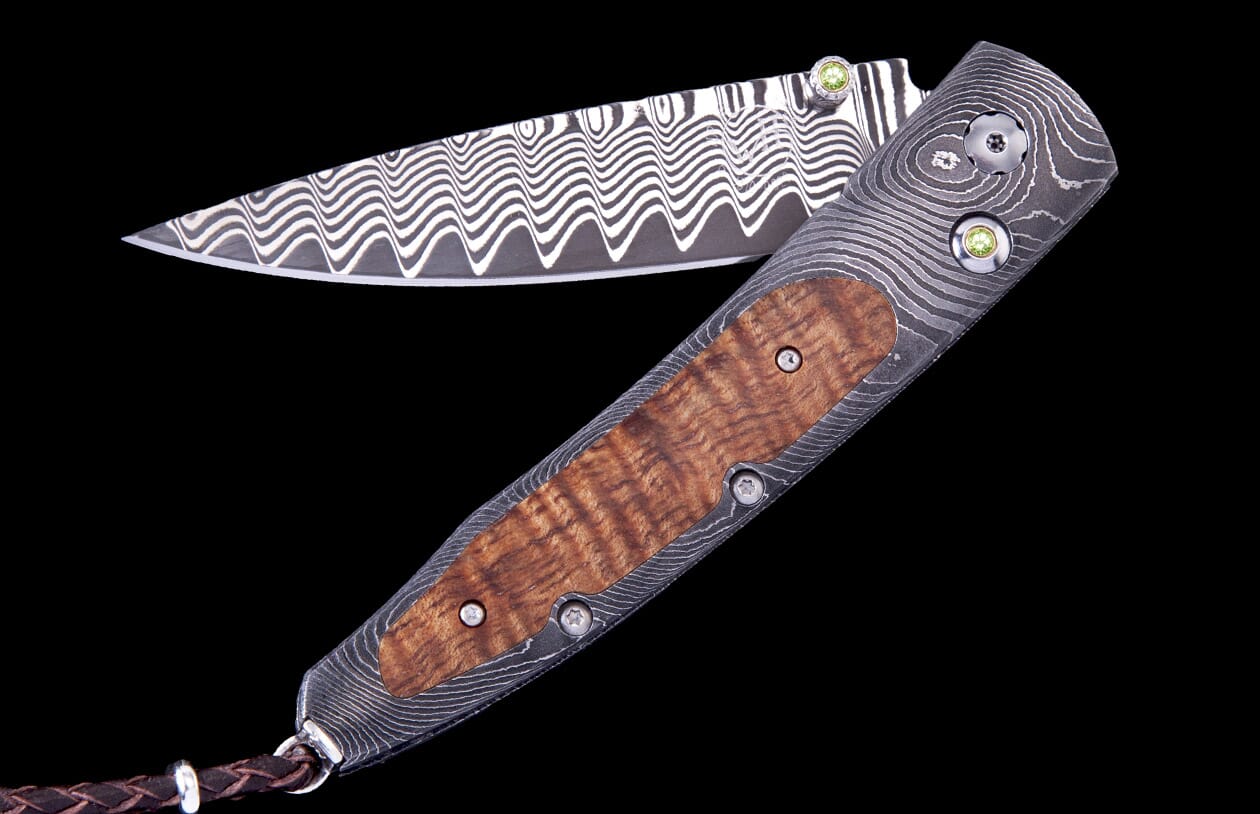Open Papakolea Exclusive Edition folding knife with wavy Damascus steel blade and handle, burled wood scales, green gemstone accents, and a brown leather lanyard.