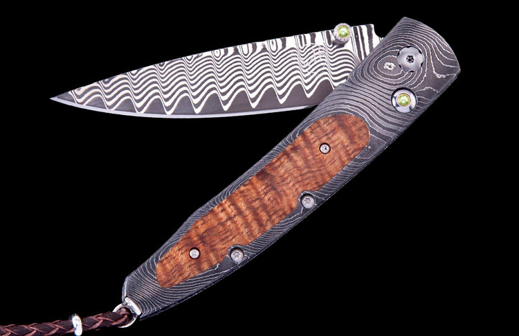 Open Papakolea Exclusive Edition folding knife with wavy Damascus steel blade and handle, burled wood scales, green gemstone accents, and a brown leather lanyard.