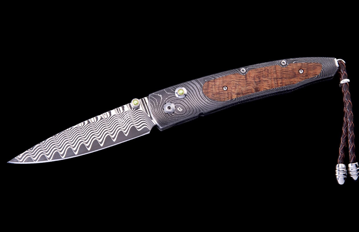 Papakolea Exclusive Edition folding knife with a wavy Damascus steel blade, wooden handle, green accents, and braided leather lanyard.