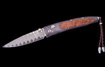 Papakolea Exclusive Edition folding knife with a wavy Damascus steel blade, wooden handle, green accents, and braided leather lanyard.
