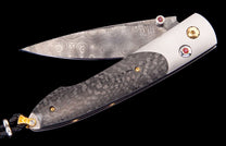 Folding knife with a swirling Damascus steel blade, dark carbon fiber handle, red gemstone accents, and a black leather lanyard.