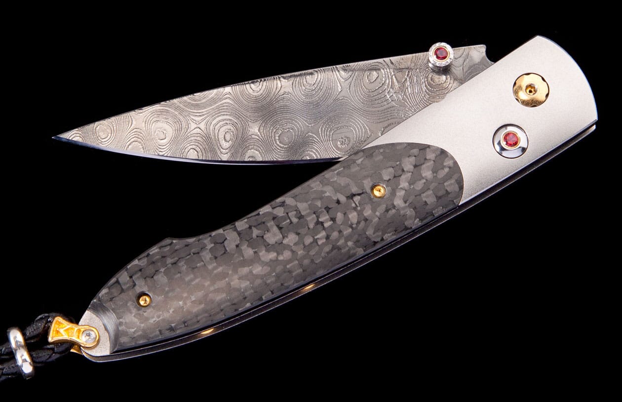 Folding knife with a swirling Damascus steel blade, dark carbon fiber handle, red gemstone accents, and a black leather lanyard.