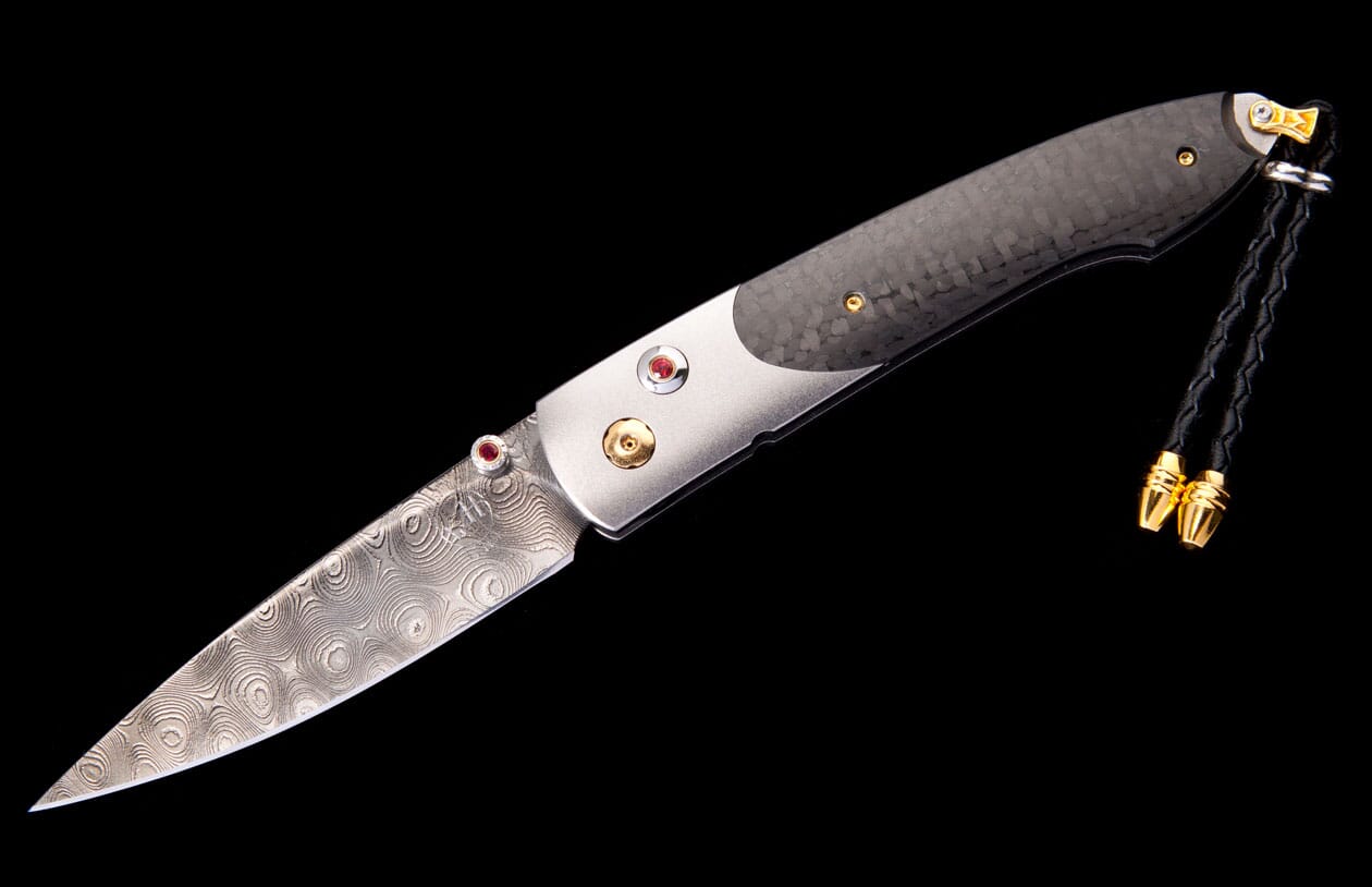 Folding knife with a textured Damascus steel blade, silver and carbon fiber handle with red and gold accents, and a black braided lanyard.