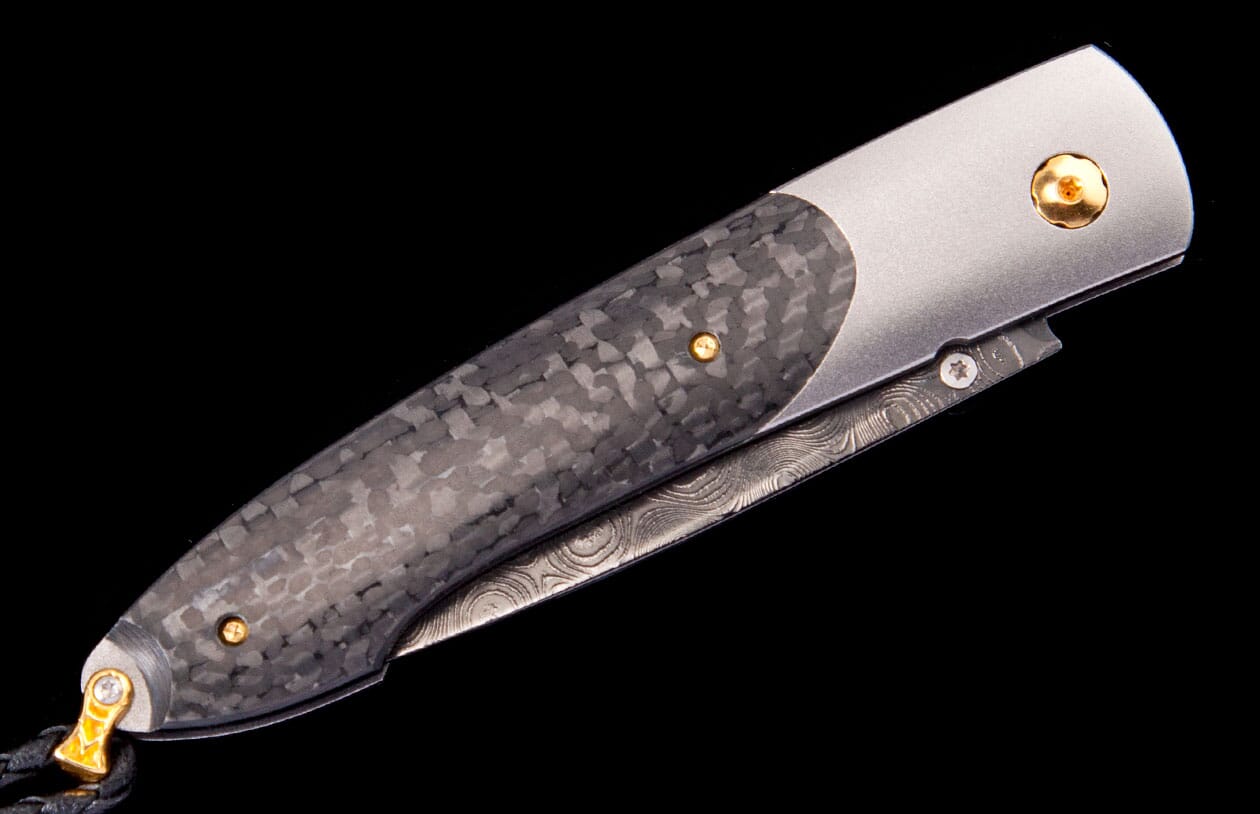 A folding knife featuring a textured carbon fiber handle, a satin metal bolster, a damascus patterned blade, and gold pins.