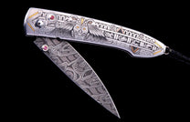 Silver-toned folding knife with intricate eagle and geometric engravings, gold accents, red gemstones, and a swirling patterned Damascus-style blade.