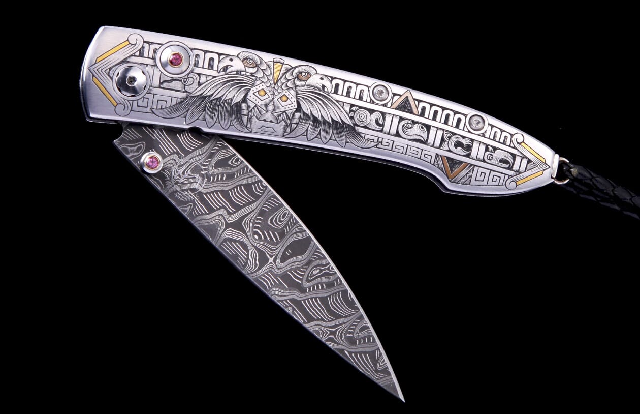Silver-toned folding knife with intricate eagle and geometric engravings, gold accents, red gemstones, and a swirling patterned Damascus-style blade.