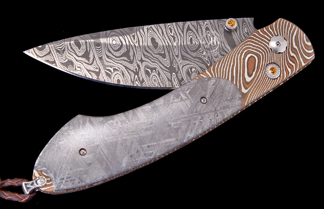 Artisanal folding knife by Carefree Buffalo Exclusive with patterned Damascus blade, striped brown and silver handle with orange studs, and a grey handle with geometric crystalline patterns.