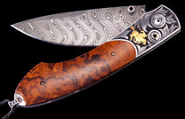Folding knife with Damascus steel blade, burled wood handle, and engraved silver bolsters featuring a gold cowboy inlay, partially open.