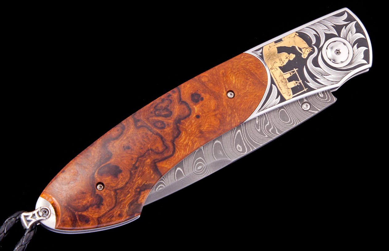Ornate folding knife with a reddish-brown burl wood handle, engraved silver bolster featuring a gold cowboy and campfire scene, and a Damascus steel blade.